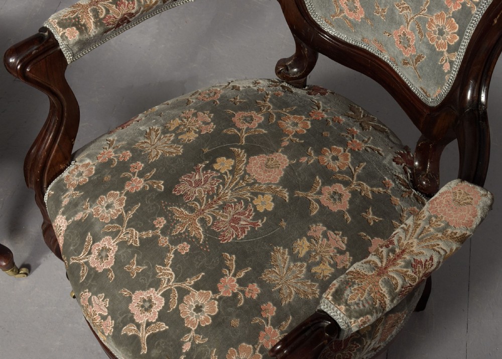 A pair of Victorian rosewood armchairs featuring carved cresting rails, padded armrests, floral patterned textile upholstery, and cabriole legs on brass castors. View III