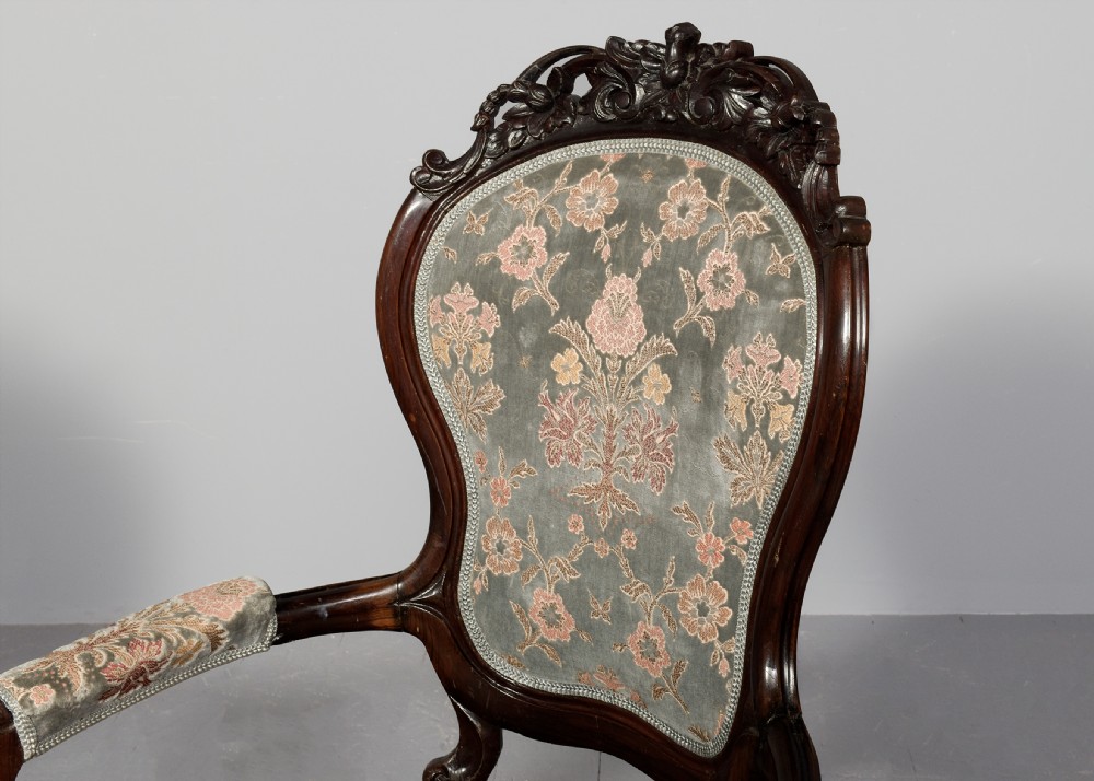A pair of Victorian rosewood armchairs featuring carved cresting rails, padded armrests, floral patterned textile upholstery, and cabriole legs on brass castors. View V