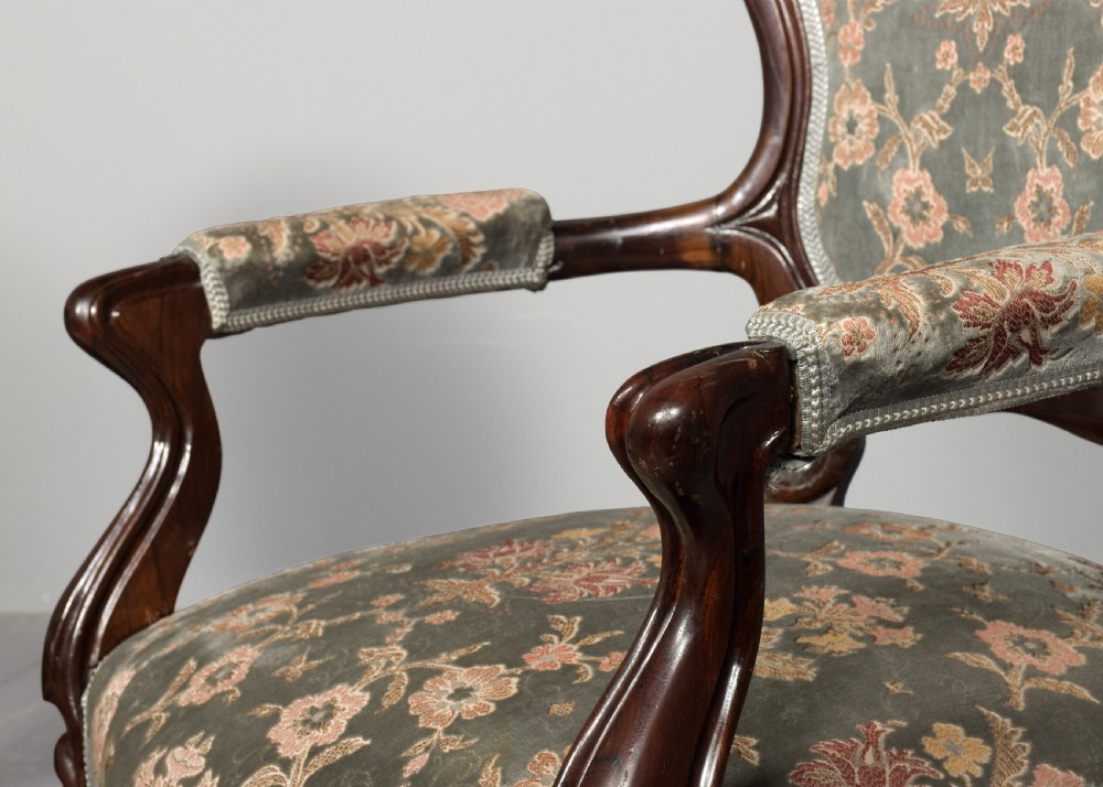 A pair of Victorian rosewood armchairs featuring carved cresting rails, padded armrests, floral patterned textile upholstery, and cabriole legs on brass castors. View VI