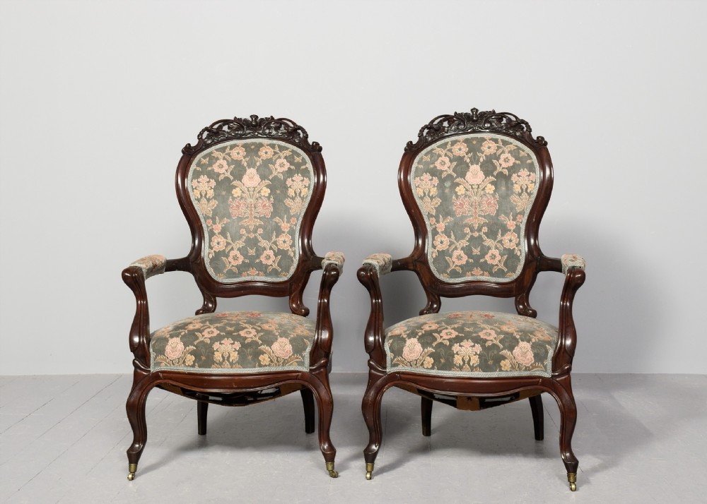 A pair of Victorian rosewood armchairs featuring carved cresting rails, padded armrests, floral patterned textile upholstery, and cabriole legs on brass castors. View VIII