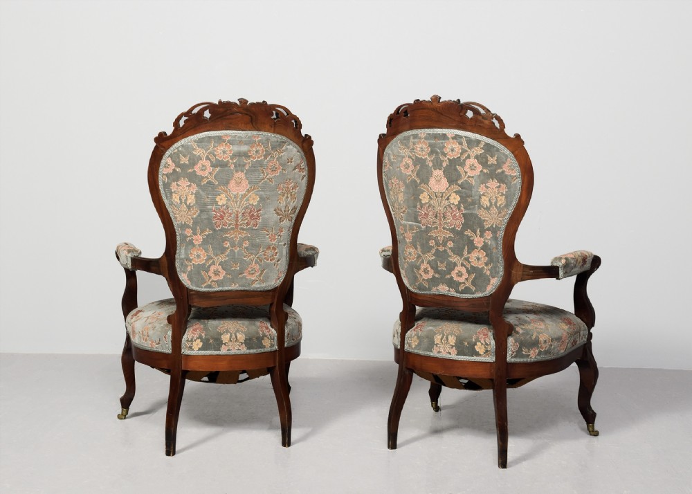A pair of Victorian rosewood armchairs featuring carved cresting rails, padded armrests, floral patterned textile upholstery, and cabriole legs on brass castors. View IX