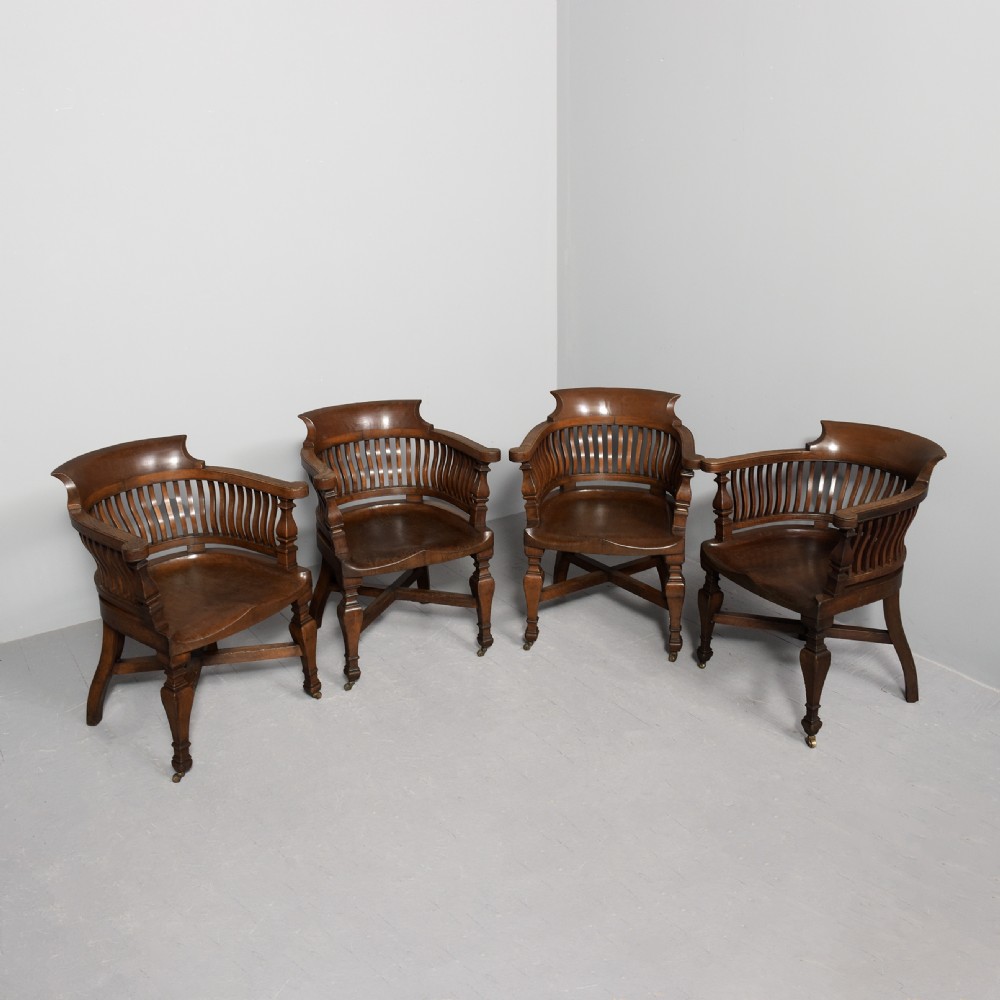 A set of four Victorian dark oak captains chairs featuring curved spindle backs, contoured seats, turned legs, and brass castors.