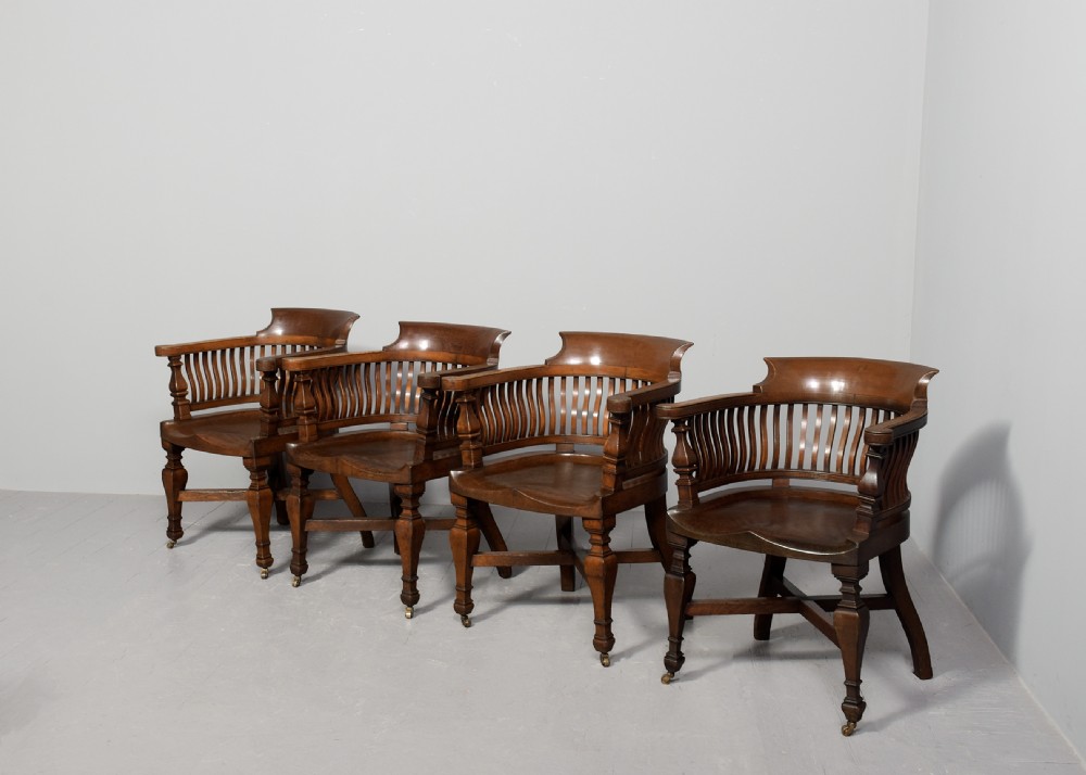A set of four Victorian dark oak captains chairs featuring curved spindle backs, contoured seats, turned legs, and brass castors. View II