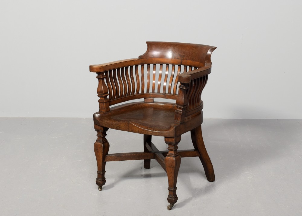 A set of four Victorian dark oak captains chairs featuring curved spindle backs, contoured seats, turned legs, and brass castors. View IV