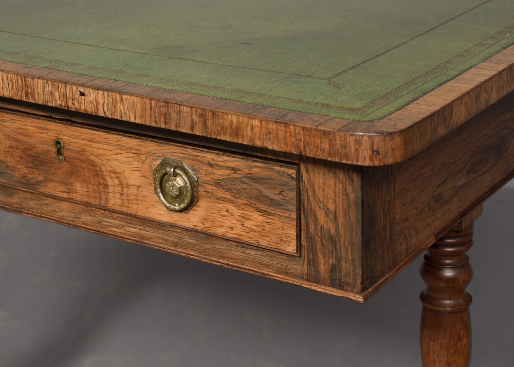 A Regency rosewood library table featuring a green leather insert top, two drawers with brass ring handles, turned supports, and splayed legs with brass castors. View IV