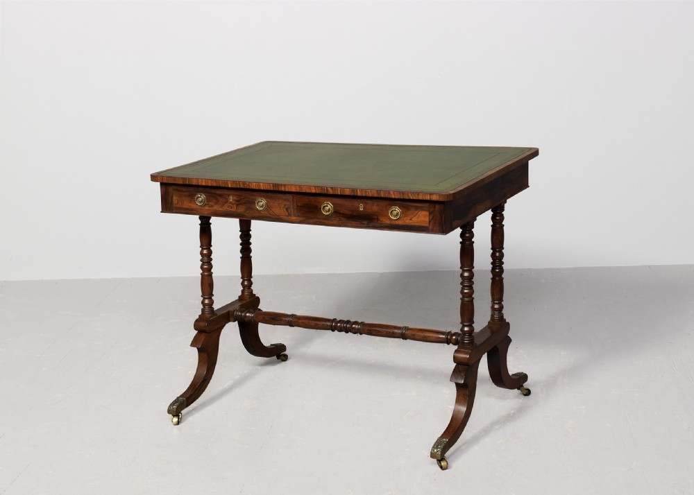A Regency rosewood library table featuring a green leather insert top, two drawers with brass ring handles, turned supports, and splayed legs with brass castors. View VI