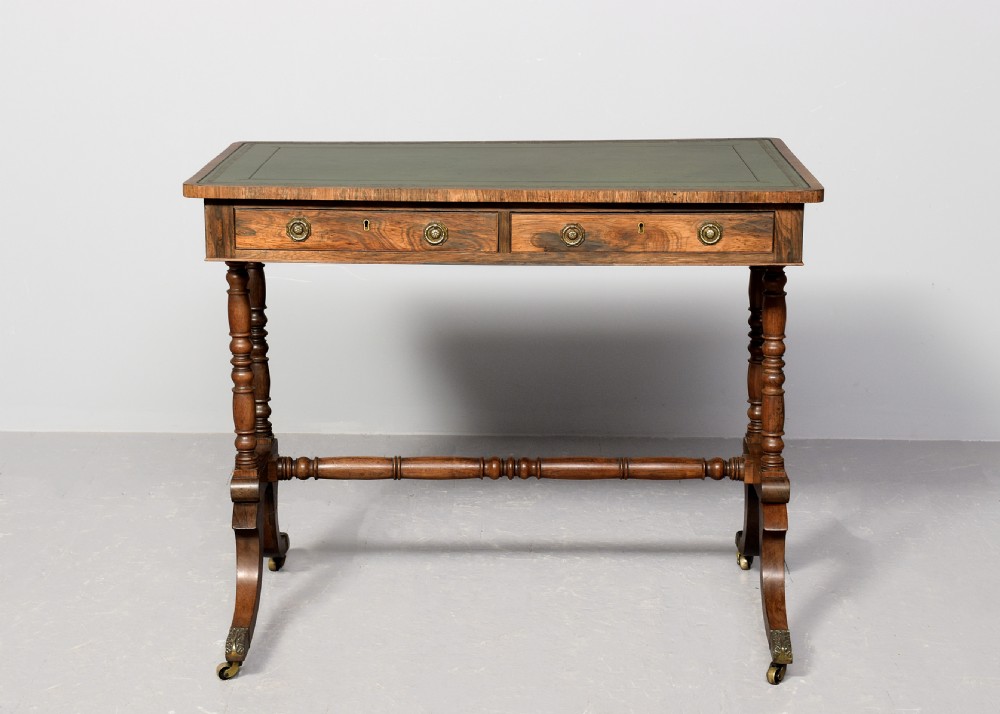 A Regency rosewood library table featuring a green leather insert top, two drawers with brass ring handles, turned supports, and splayed legs with brass castors. View VII