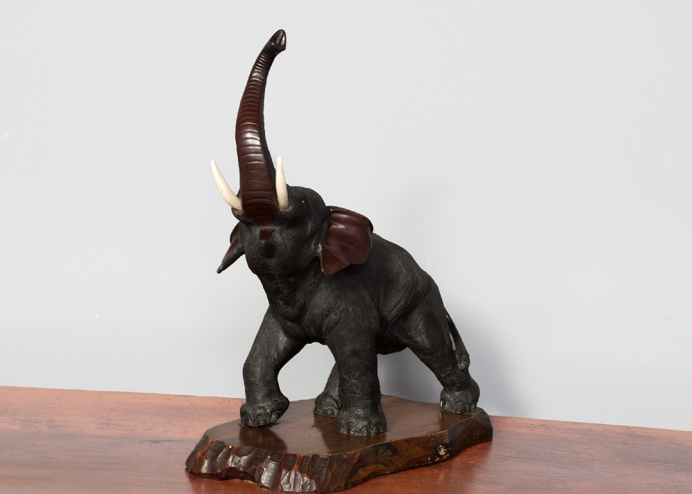 A dark patinated Japanese bronze elephant sculpture with raised trunk and white tusks, mounted on a naturalistic cut wood base. View VI