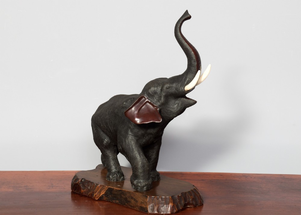 A dark patinated Japanese bronze elephant sculpture with raised trunk and white tusks, mounted on a naturalistic cut wood base. View VII