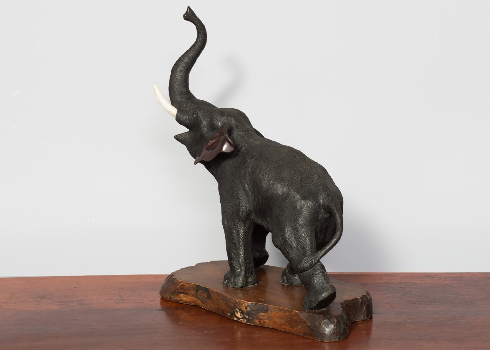 A dark patinated Japanese bronze elephant sculpture with raised trunk and white tusks, mounted on a naturalistic cut wood base. View VIII