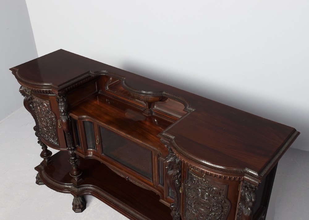 A late Victorian dark mahogany sideboard featuring detailed carved relief cupboard doors, a central glazed cabinet, mirrored back panel, and carved lion paw feet. View IX