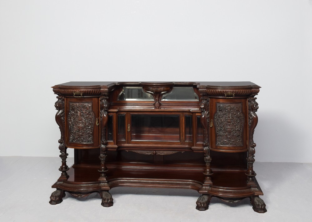 A late Victorian dark mahogany sideboard featuring detailed carved relief cupboard doors, a central glazed cabinet, mirrored back panel, and carved lion paw feet. View X