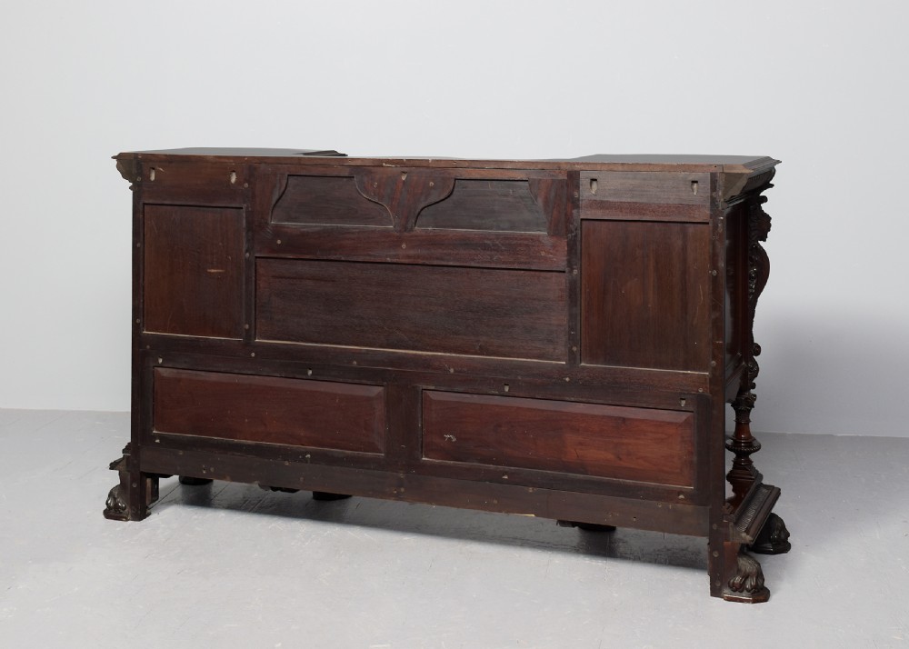 A late Victorian dark mahogany sideboard featuring detailed carved relief cupboard doors, a central glazed cabinet, mirrored back panel, and carved lion paw feet. View XI