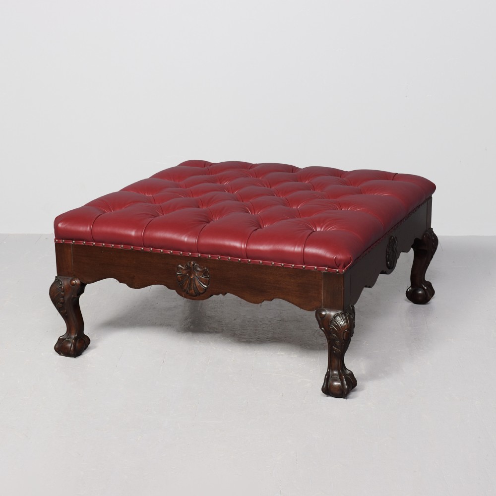 A large square mahogany footstool featuring deep-buttoned burgundy leather upholstery, brass studding, and carved cabriole legs with ball-and-claw feet.