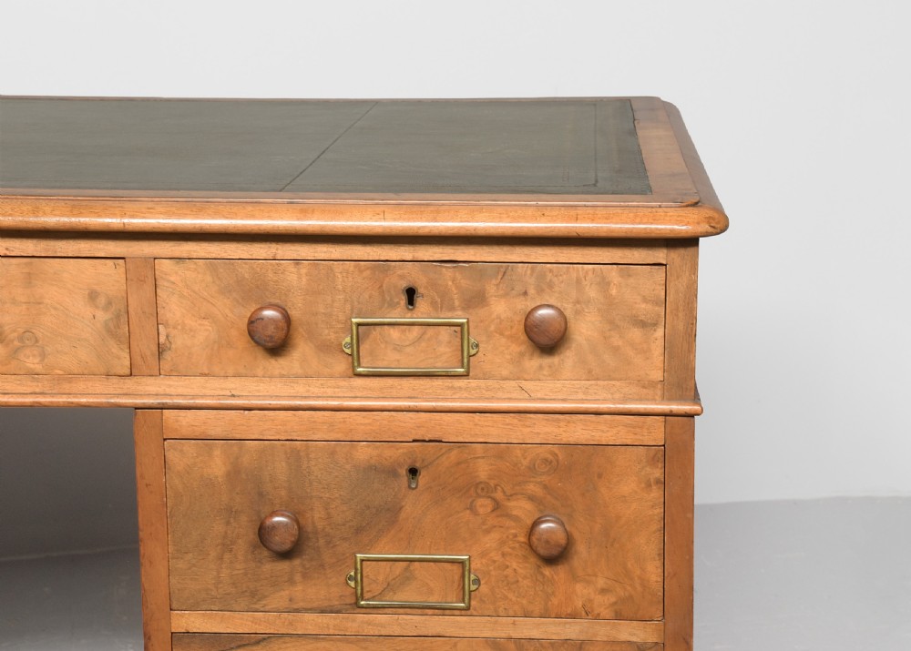 Victorian figured walnut pedestal desk featuring a green gilt-tooled leather writing surface, multiple drawers with turned wooden knobs, brass card handles, and plinth bases. View V