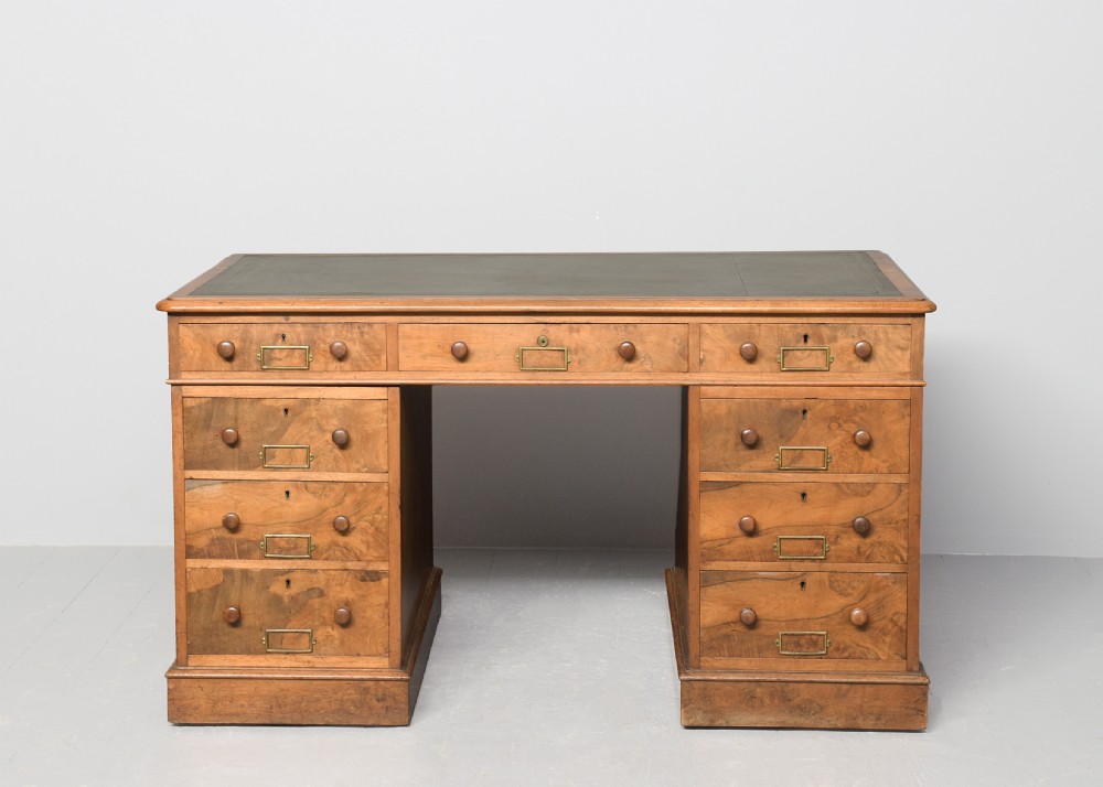 Victorian figured walnut pedestal desk featuring a green gilt-tooled leather writing surface, multiple drawers with turned wooden knobs, brass card handles, and plinth bases. View VI
