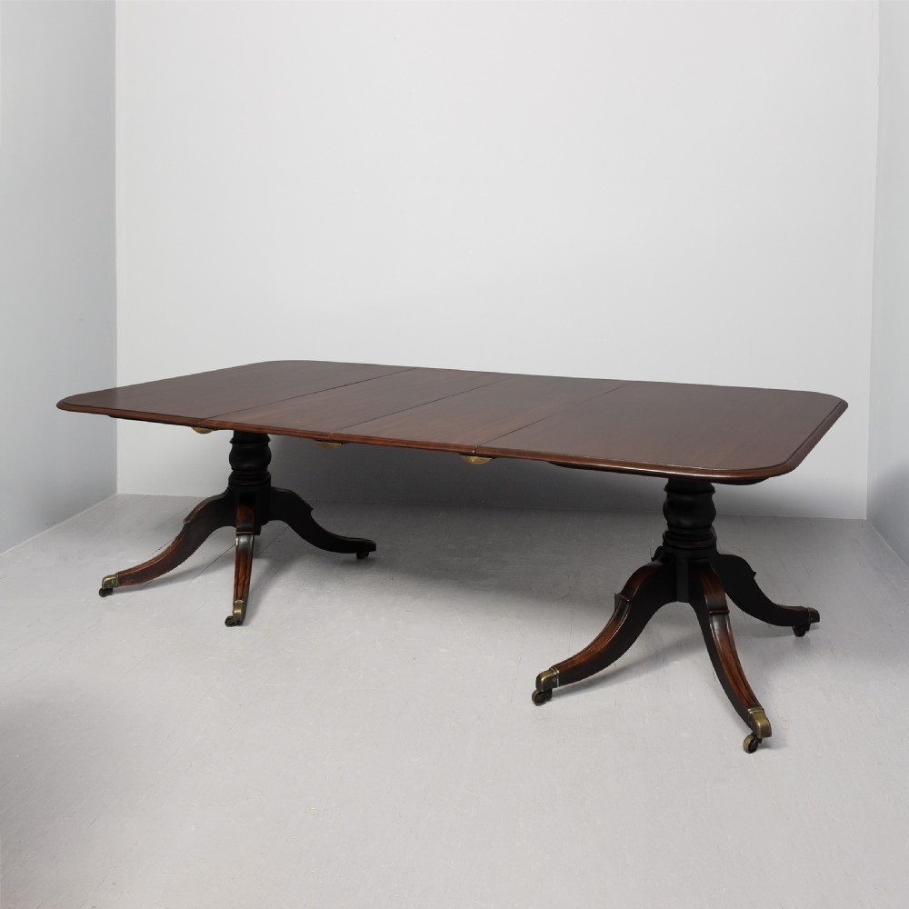An extended George IV mahogany dining table featuring two turned pedestal supports with splayed legs, brass castors, and two removable leaf sections.