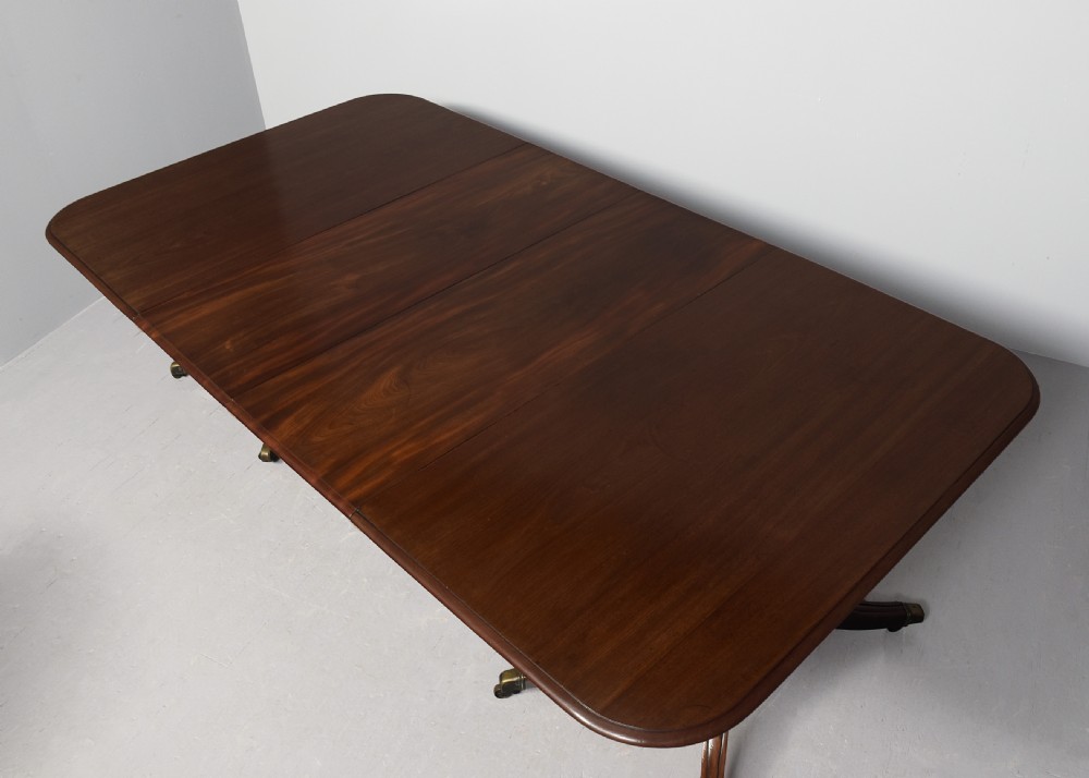 An extended George IV mahogany dining table featuring two turned pedestal supports with splayed legs, brass castors, and two removable leaf sections. View II