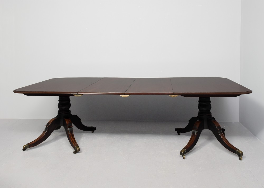 An extended George IV mahogany dining table featuring two turned pedestal supports with splayed legs, brass castors, and two removable leaf sections. View III