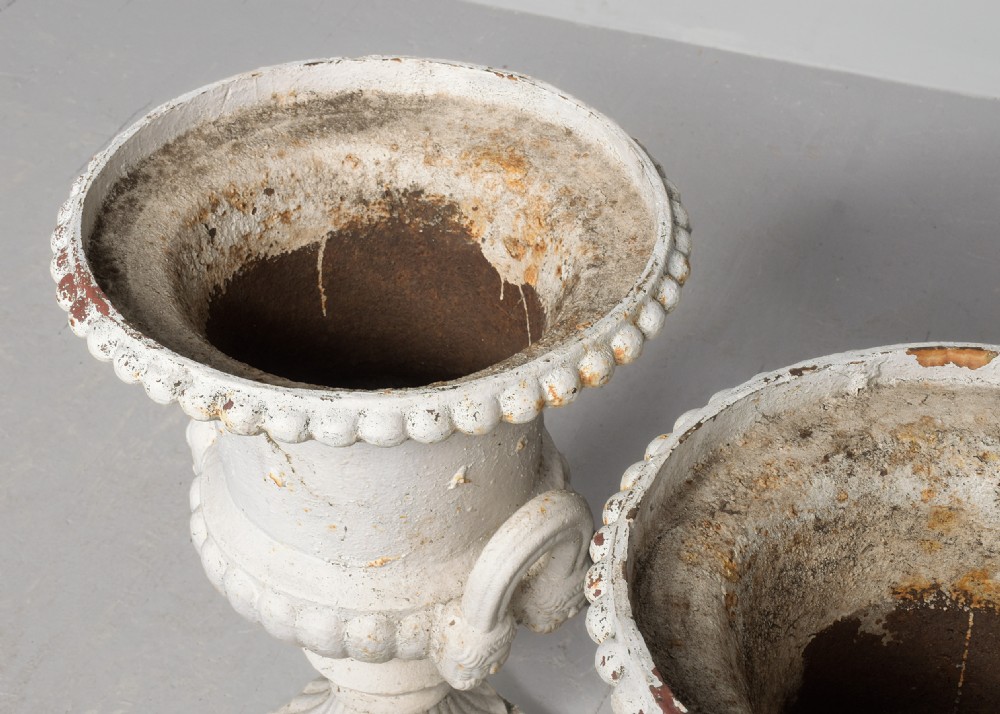 A pair of classical off-white painted cast iron Campagna urns featuring gadrooned rims, looped handles, and square pedestal bases displaying surface rust and patination. View II