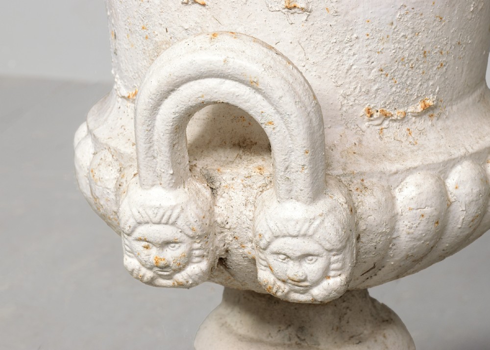 A pair of classical off-white painted cast iron Campagna urns featuring gadrooned rims, looped handles, and square pedestal bases displaying surface rust and patination. View V