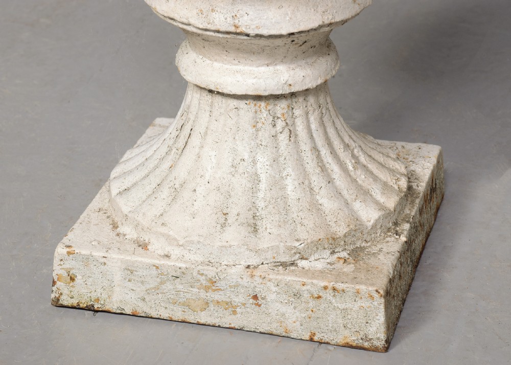 A pair of classical off-white painted cast iron Campagna urns featuring gadrooned rims, looped handles, and square pedestal bases displaying surface rust and patination. View VI