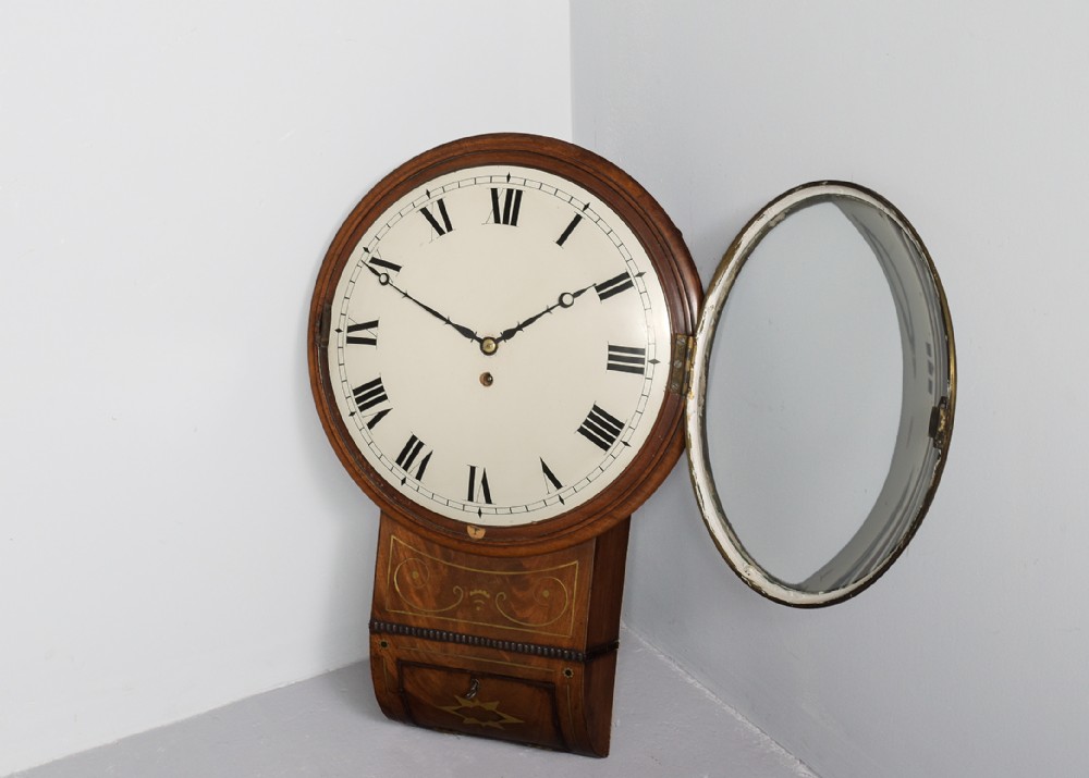 An antique mahogany drop dial fusee wall clock featuring brass inlay decoration, a circular white dial with black Roman numerals, and an open hinged brass bezel.