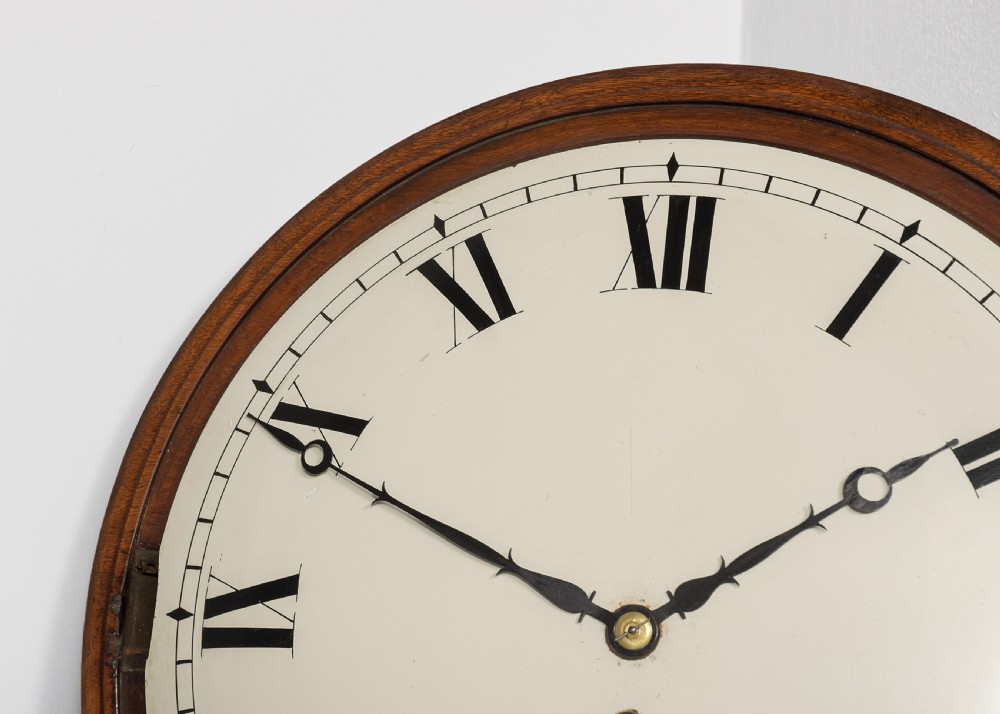 An antique mahogany drop dial fusee wall clock featuring brass inlay decoration, a circular white dial with black Roman numerals, and an open hinged brass bezel. View III