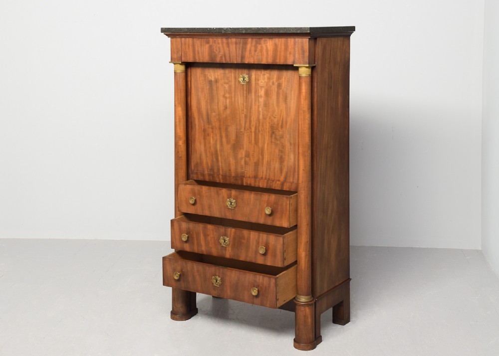 A tall mahogany secretaire abattant featuring a dark marble top, a drop-front writing panel flanked by turned columns, and three lower drawers with brass handles. View IV