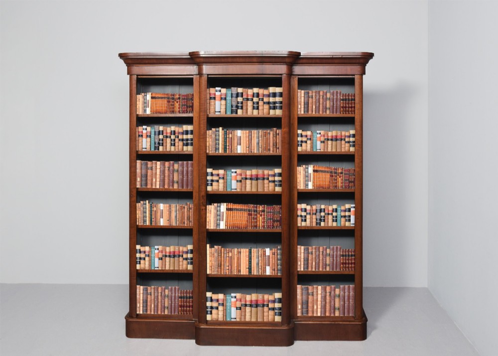 A large mid-Victorian three-section open mahogany bookcase with a breakfront centre, filled with rows of leather-bound books.