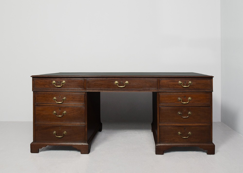 A large George III mahogany partners desk featuring a green leather writing surface, dual pedestals with brass swan-neck handles, and bracket feet. View VI