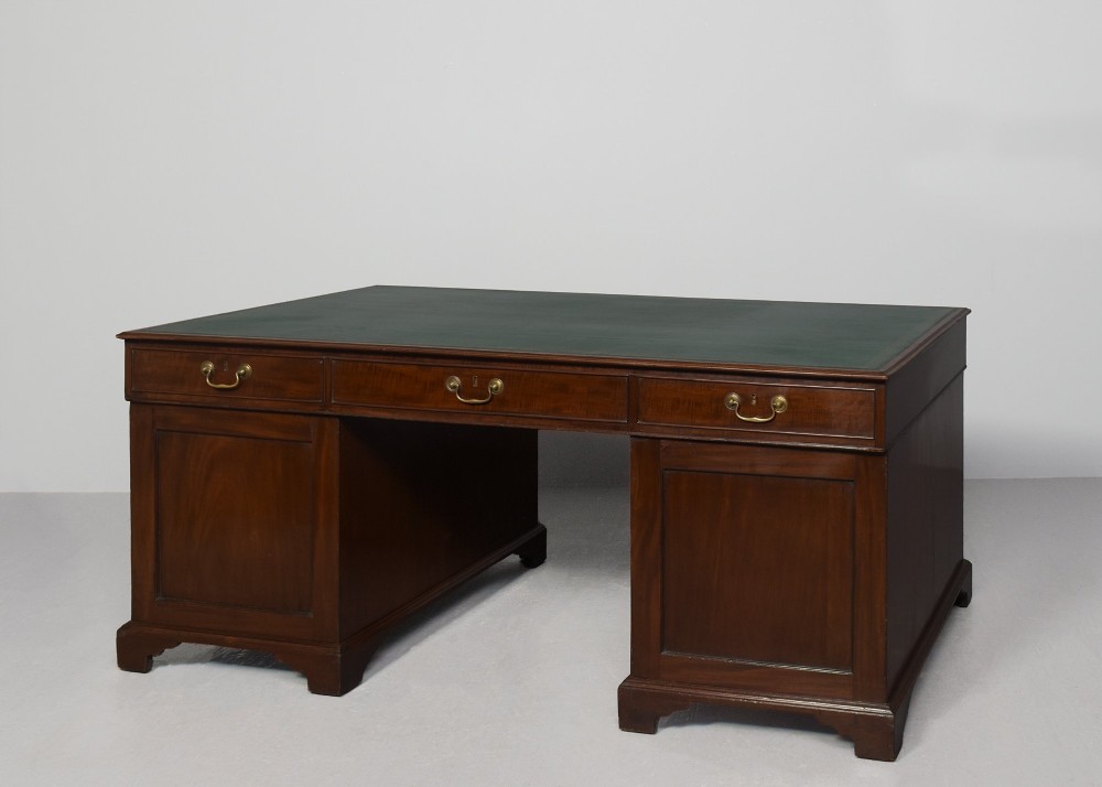 A large George III mahogany partners desk featuring a green leather writing surface, dual pedestals with brass swan-neck handles, and bracket feet. View IX