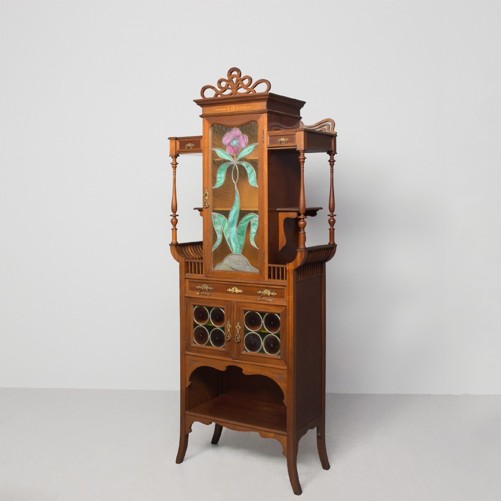 An Art Nouveau mahogany display cabinet featuring a central stained glass floral panel door, open side shelves with spindle supports, brass hardware, and round bullseye glass lower doors.