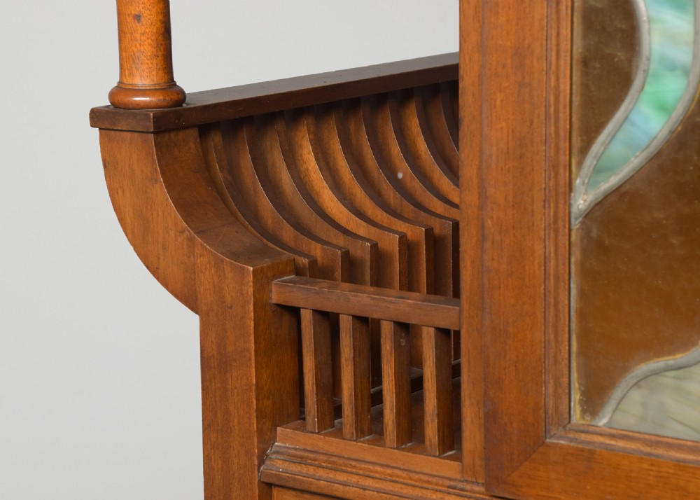 An Art Nouveau mahogany display cabinet featuring a central stained glass floral panel door, open side shelves with spindle supports, brass hardware, and round bullseye glass lower doors. View IV