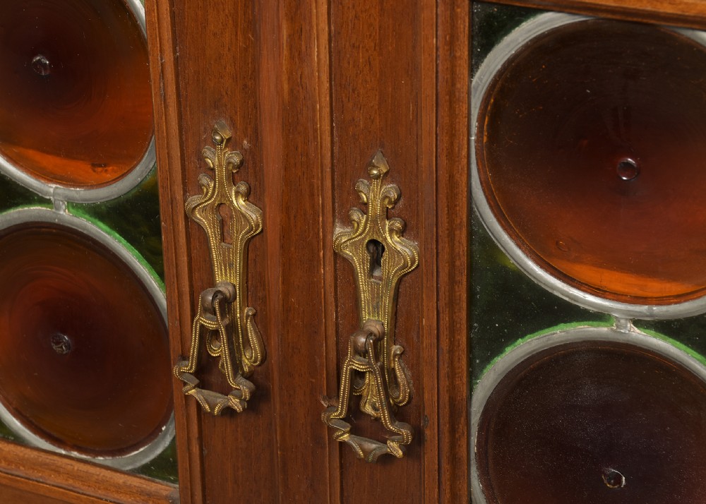 An Art Nouveau mahogany display cabinet featuring a central stained glass floral panel door, open side shelves with spindle supports, brass hardware, and round bullseye glass lower doors. View V