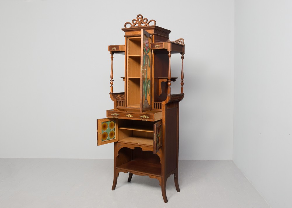 An Art Nouveau mahogany display cabinet featuring a central stained glass floral panel door, open side shelves with spindle supports, brass hardware, and round bullseye glass lower doors. View VII