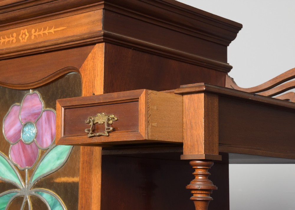 An Art Nouveau mahogany display cabinet featuring a central stained glass floral panel door, open side shelves with spindle supports, brass hardware, and round bullseye glass lower doors. View IX