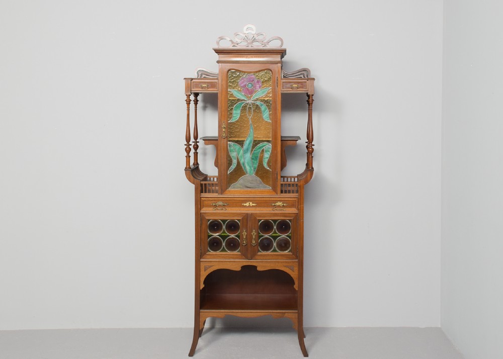 An Art Nouveau mahogany display cabinet featuring a central stained glass floral panel door, open side shelves with spindle supports, brass hardware, and round bullseye glass lower doors. View X