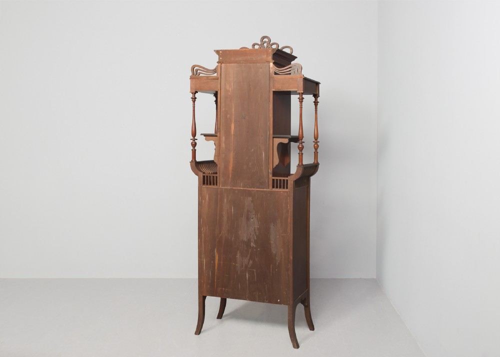 An Art Nouveau mahogany display cabinet featuring a central stained glass floral panel door, open side shelves with spindle supports, brass hardware, and round bullseye glass lower doors. View XI