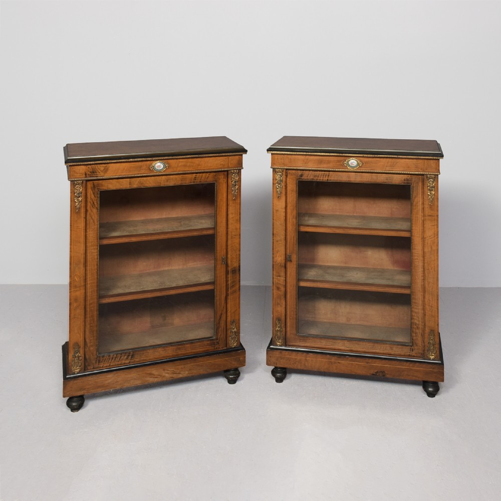 A pair of Victorian burr walnut pier cabinets with single glazed doors, ebonised trim, ormolu corner mounts, and oval porcelain plaques.