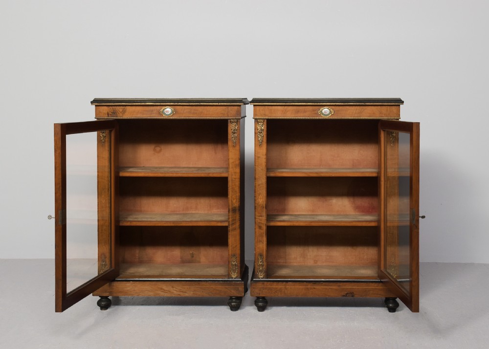 A pair of Victorian burr walnut pier cabinets with single glazed doors, ebonised trim, ormolu corner mounts, and oval porcelain plaques. View V