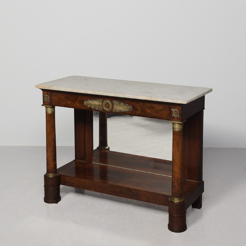 An early nineteenth-century French Empire mahogany console table featuring a light marble top, gilt brass column mounts, a mirrored back panel, and a lower shelf.