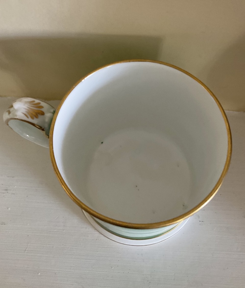 A 19th-century porcelain mug featuring a green ground, a hand-painted floral cartouche surrounded by gilded scrolls, and a white stepped base. View IV