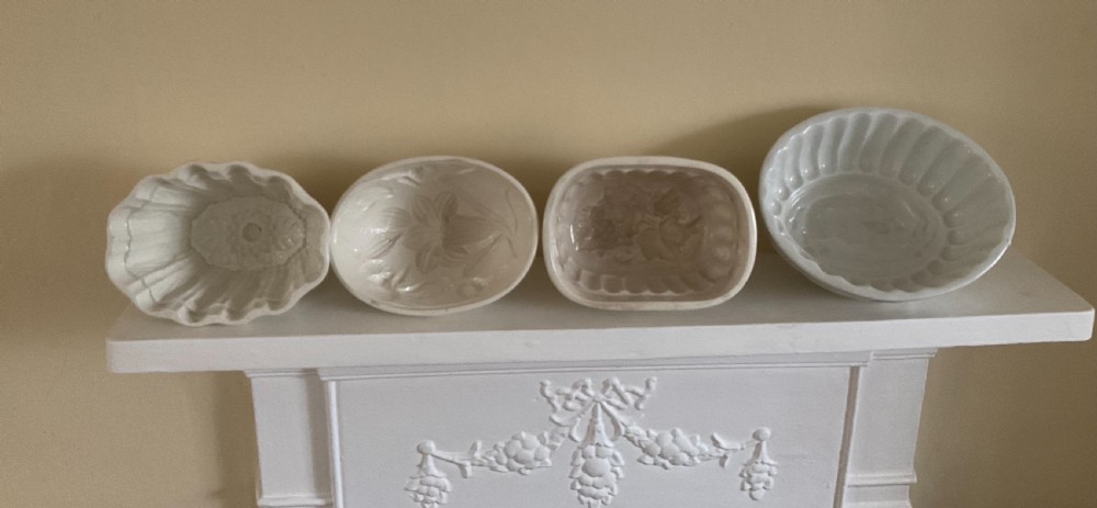 A row of four white glazed earthenware jelly moulds displaying distinct interior relief motifs including grapevines, flowers, and a boar.