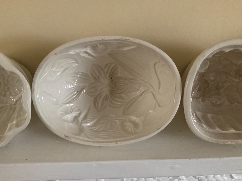 A row of four white glazed earthenware jelly moulds displaying distinct interior relief motifs including grapevines, flowers, and a boar. View III