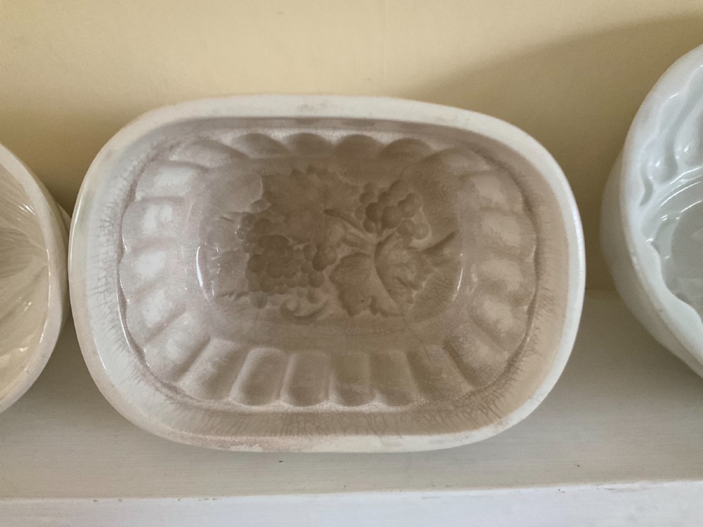 A row of four white glazed earthenware jelly moulds displaying distinct interior relief motifs including grapevines, flowers, and a boar. View IV