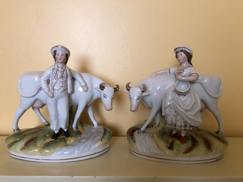 A pair of Victorian Staffordshire ceramic figures featuring a boy standing beside a white cow on one base, and a milkmaid with a pail beside a cow on the other base.