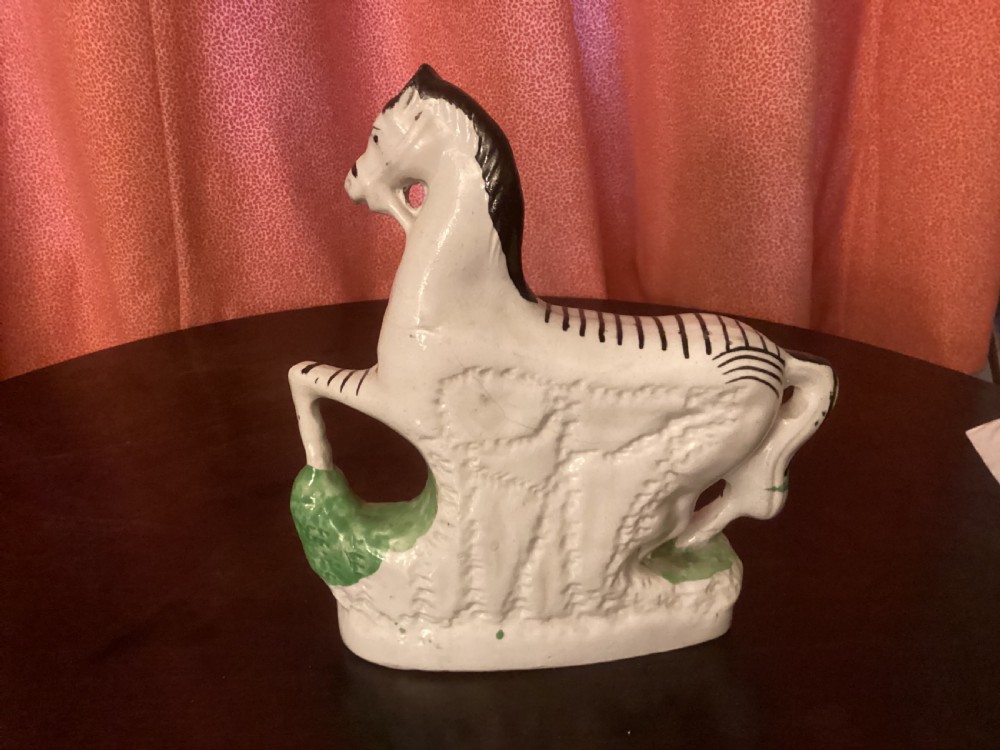 A 19th-century Staffordshire earthenware ceramic figure of a zebra with hand-painted black stripes standing on a green grass-like base. View II