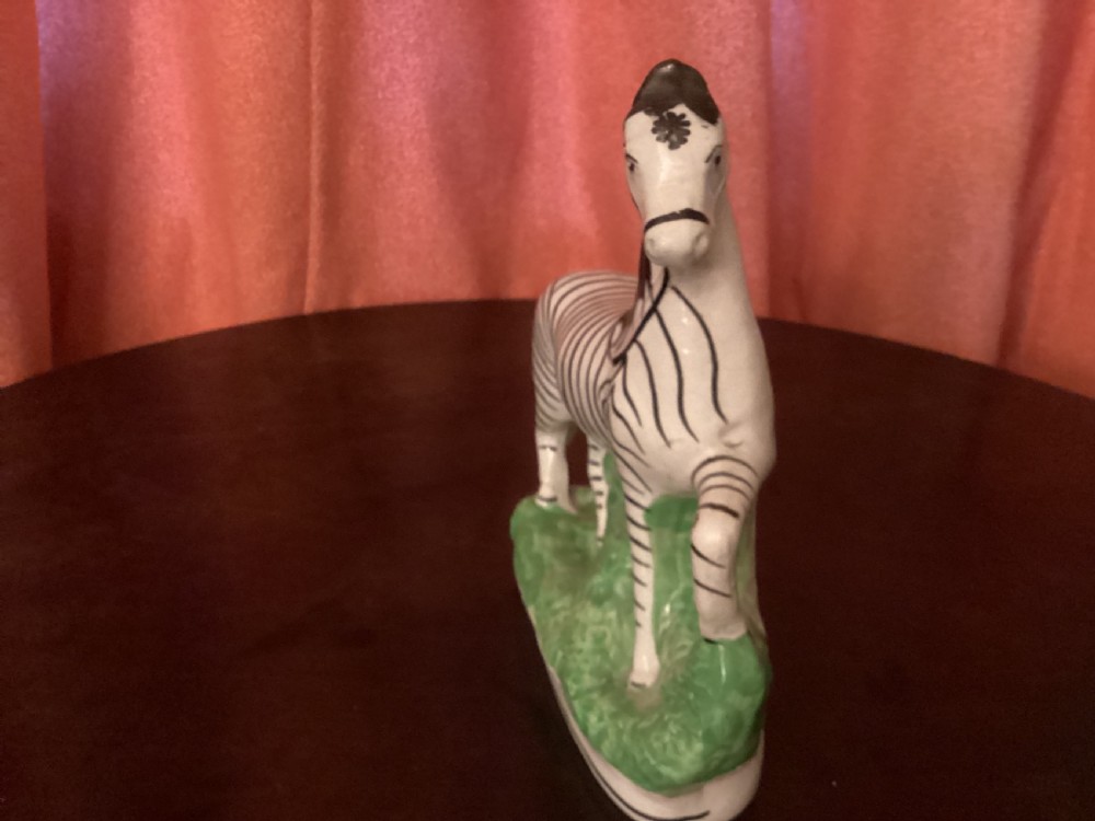 A 19th-century Staffordshire earthenware ceramic figure of a zebra with hand-painted black stripes standing on a green grass-like base. View IV
