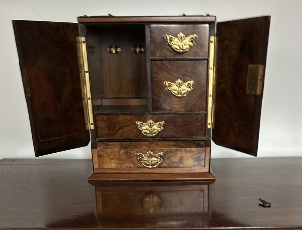 A Victorian burr walnut cigar cabinet featuring two doors adorned with pierced brass corner brackets and a central lock mechanism above a lower drawer. View IV